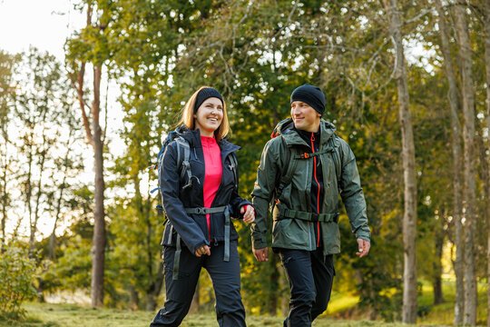 A cheerful man and woman wearing hiking gear and backpacks walk together through a forest trail, enjoying a sunny day outdoors in early autumn