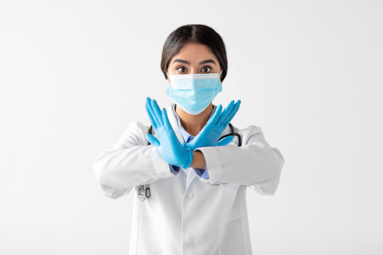A serious young Indian lady doctor in a uniform, gloves, and a protective mask crosses her arms and makes a no sign with her hands. She emphasizes the importance of health measures.