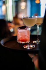 A bartender carries a tray with three cocktails, two in coupe glasses and one in an old-fashioned glass, in a dimly lit bar or restaurant setting