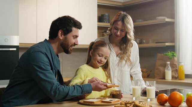 Happy Caucasian family at kitchen at home preparing breakfast together mother hold glasses with milk little daughter child girl prepare toasts bread with chocolate pasta with father help food delivery