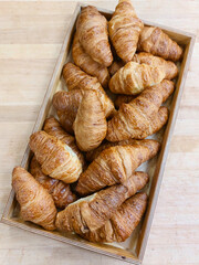 Golden crispy croissants arranged in a wooden box on a light wooden background. Warm natural light highlighting texture and fresh pastry crust. Perfect image for bakery, breakfast or cafe concept.