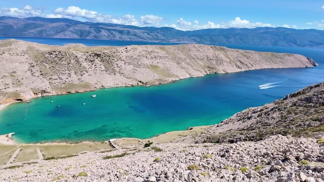 Aerial view of turquoise rocky coastline with boats and waves, sunny skies, and scenic landscape. Vela Luka beach, Krk island, Croatia