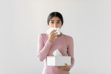 A shocked young woman struggles with flu symptoms, blowing her nose and holding a box of tissues. She appears upset in a simple gray background, highlighting her health issues and discomfort.