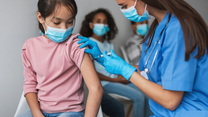 A nurse is giving a Covid-19 vaccine to a young Asian girl in a contemporary clinic. The girl is...