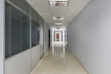 A long corridor stretches through a modern office space. Offices line the sides, and natural light brightens the floor. The walls are painted light grey