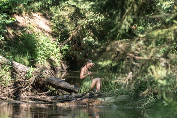 Young woman with short hair, dressed in a green wrap, sits gracefully by a serene riverbank surrounded by lush greenery, embodying tranquility and connection with nature