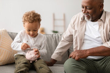 African American grandfather and his grandson sit together on a couch. The grandson is putting coins into a piggybank, learning about saving money and financial responsibility.