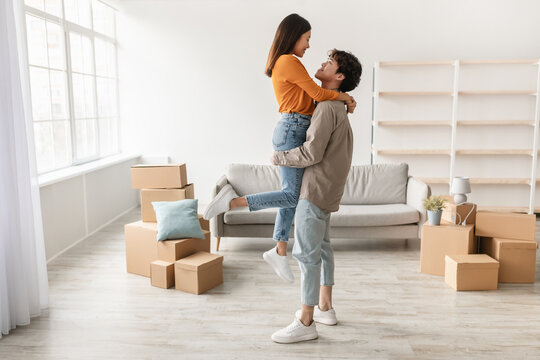 Young Asian couple happily embraces while moving into their new house. The man lifts his girlfriend in an empty room filled with cardboard boxes, symbolizing a fresh start.