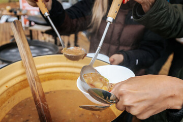 A steaming bowl of hearty soup is served from a large pot outdoors, offering warmth and comfort during a cozy autumn gathering.