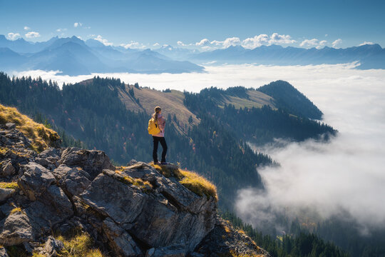 Woman with yellow backpack standing on mountain ridge above a sea of clouds in the Swiss Alps. Scenic landscape with girl, blue sky, distant peaks, and bright sunlight in autumn. Hiking in Switzerland
