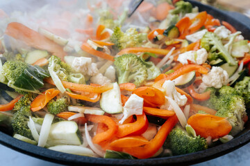A vibrant mix of steaming stir-fried vegetables including carrots, broccoli, zucchini, cauliflower, and bell peppers, cooking in a large black pan outdoors.