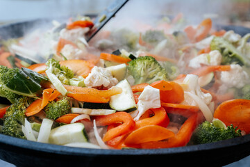 A vibrant mix of steaming stir-fried vegetables including carrots, broccoli, zucchini, cauliflower, and bell peppers, cooking in a large black pan outdoors.