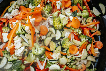 A vibrant mix of steaming stir-fried vegetables including carrots, broccoli, zucchini, cauliflower, and bell peppers, cooking in a large black pan outdoors.