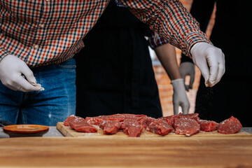 A person wearing gloves and a plaid shirt seasons raw steaks with ground pepper on a wooden board during an outdoor cooking demonstration.