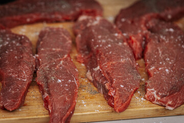 Close-up of raw beef steaks seasoned with salt and pepper, laid out on a wooden cutting board, ready for grilling at an outdoor cooking event.