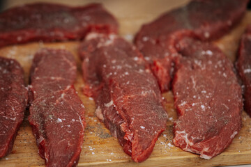 Close-up of raw beef steaks seasoned with salt and pepper, laid out on a wooden cutting board, ready for grilling at an outdoor cooking event.