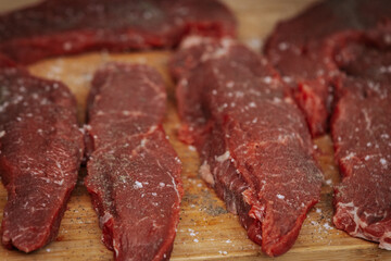 Close-up of raw beef steaks seasoned with salt and pepper, laid out on a wooden cutting board, ready for grilling at an outdoor cooking event.