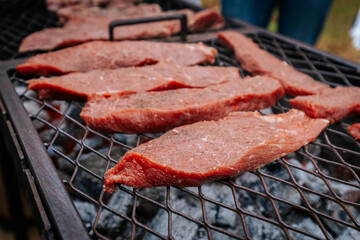 Close-up of raw beef steaks sizzling on a grill over hot coals, with visible seasoning and sear marks beginning to form.