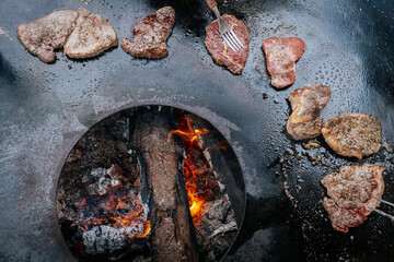 Raw steaks sizzle in a circle on a hot flat grill surrounding an open wood fire, capturing a...