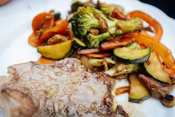 Close-up of a juicy grilled steak served with colorful sautéed vegetables and beans, glistening with herbs and oil on a white plate.