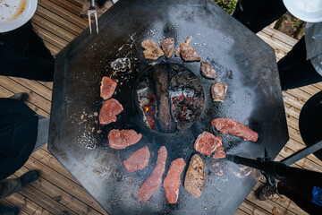 Raw steaks sizzle in a circle on a hot flat grill surrounding an open wood fire, capturing a...