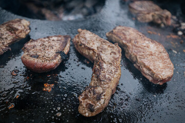 Raw steaks sizzle in a circle on a hot flat grill surrounding an open wood fire, capturing a dramatic top-down view of live-fire cooking in progress.