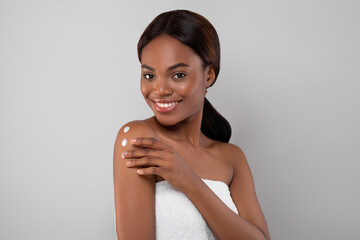 A woman is applying lotion on her shoulder with a joyful smile. She is wrapped in a towel and...