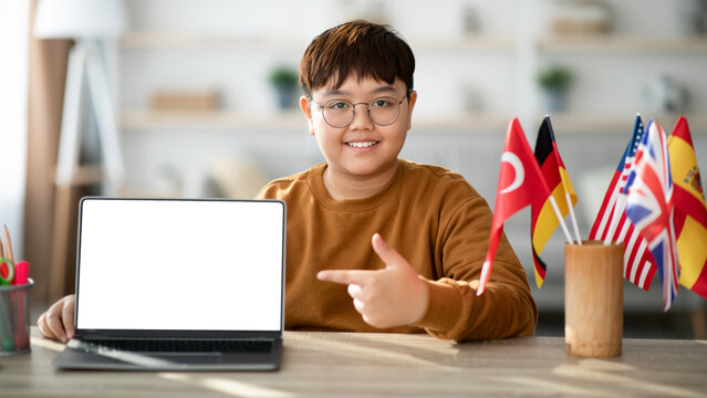 A positive chubby asian boy sits at a desk, smiling and pointing at a modern laptop with a blank screen. He is engaged in an online class, studying a foreign language. Flags are displayed.
