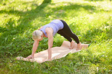 Middle-aged woman exercises and does core strengthening exercises. Elderly woman practices fitness and yoga outside in park on sunny day, laying out fitness mat on grass.