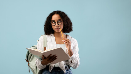 A thoughtful black female student reads a textbook and ponders her assignment. She stands against a blue studio background, holding a pencil and wearing a backpack, focused on solving her problem.