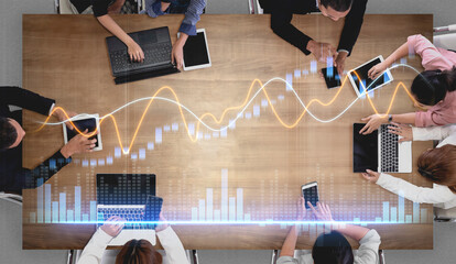A top view of a diverse group of professionals engaged in a collaborative meeting, analyzing data trends on digital devices, showcasing teamwork and innovation. Scalp