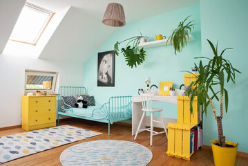 Room child in the attic with window, carpet in modern design on the wooden floor, turquoise wall and yellow furniture. Cozy interior full of color with desk, bed and plant.