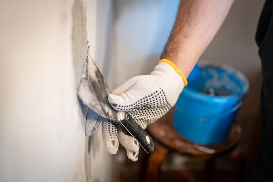 Close-up of a worker's gloved hand using a small putty knife to press thick patching compound firmly into a hole in the interior wall.