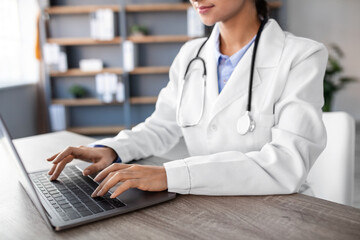 A young Hindu woman doctor wearing a white coat and stethoscope types on a laptop in her office....