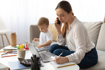 A mother balances work and parenting in a bright living room. She is on the phone, using a laptop, while her young child plays nearby, creating a warm and productive atmosphere.