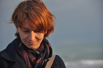 Young woman with short red hair smiles softly while standing on the beach during a cloudy day