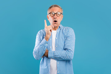 Mature man in glasses looks surprised and joyful, pointing finger up as he thinks of a great idea. He stands against a bright blue wall, showing excitement and inspiration. © Prostock-studio