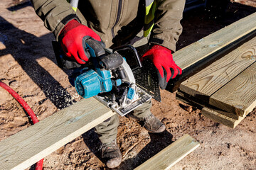 Carpenter cutting wooden plank with circular saw outdoors, Worker wearing red gloves uses a circular saw to cut treated wooden boards on a construction site during a woodworking project.