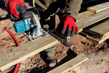 Carpenter cutting wooden plank with circular saw outdoors, Worker wearing red gloves uses a circular saw to cut treated wooden boards on a construction site during a woodworking project.