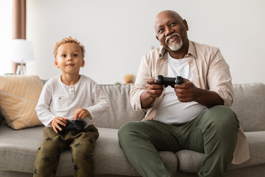 Cheerful grandfather and his grandson sit on the couch playing video games at home. They share laughter and enjoyment while holding gamepad controllers. It's a perfect weekend bonding moment.