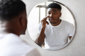 In a stylish bathroom, an African American man applies cream moisturizer under his eyes while...