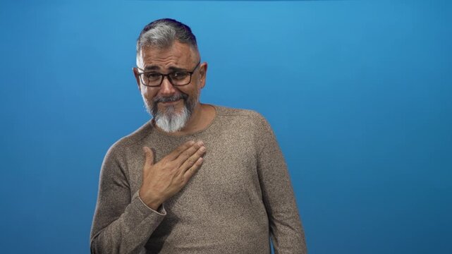 Middle age man smiling with hand on chest in studio portrait against solid blue backdrop; gratitude modesty reflection.