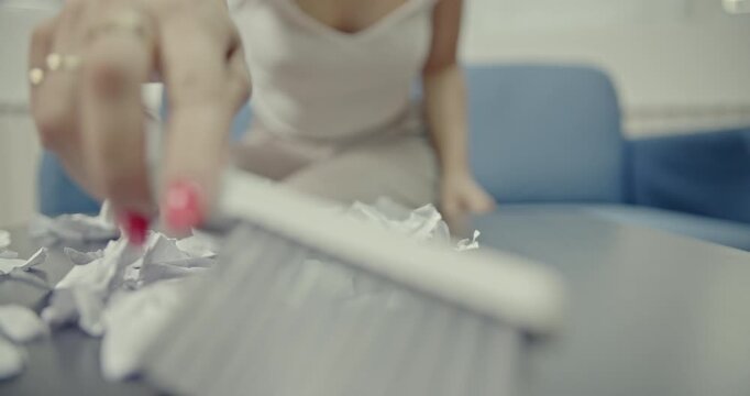 A woman sweeps crumpled paper from a table into a dustpan with a brush, featuring crisp and satisfying cleaning sounds. This video captures the detailed act of tidying up and organizing a space.