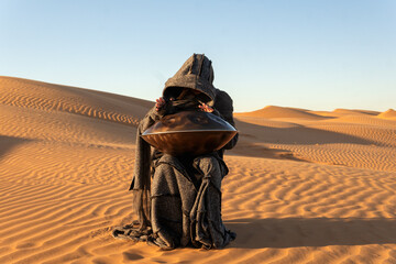 A girl in a long coat plays the handpan against the backdrop of dunes