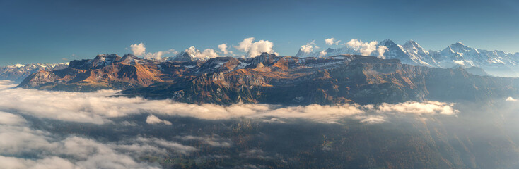 Panoramic aerial view of the Swiss Alps with snow-capped peaks and low clouds at sunset. Bright evening light and clear blue sky over rugged alpine mountains in Switzerland. Top view. Landscape