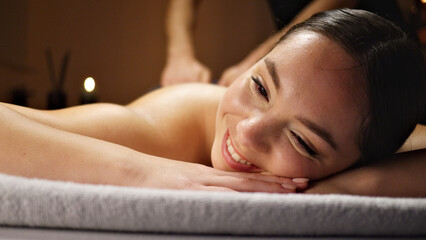 Obraz premium A woman smiles serenely on a massage table as a therapist works on her back. The warm, inviting setting with soft lighting and candles creates a relaxing atmosphere, emphasizing wellness, self-care.