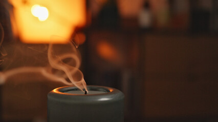 A lit incense burner releases delicate smoke spirals under warm, ambient lighting, creating a serene, contemplative atmosphere. Ideal for relaxation or meditation, the soft glow.