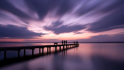 A captivating view of a wooden pier stretching into tranquil waters beneath a dramatic sky filled with long exposure clouds. A group of people stand at the end, adding a sense of scale and serenity.