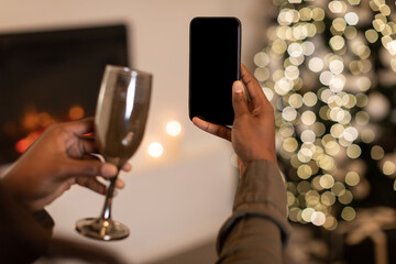 An unrecognizable African American lady celebrates Christmas at home by video calling on her smartphone. She clinks a glass, enjoying the festive moment and cozy atmosphere.