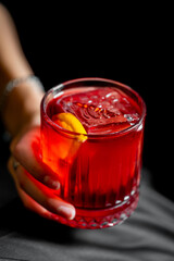 A vibrant red Negroni cocktail with a large ice cube and orange peel, held in a fluted rocks glass against a dark background, in a close-up shot.